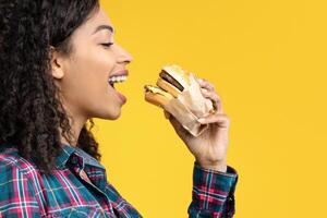 Time For Snack. Side View Portrait Of Hungry Black Lady Eating Hamburger. African American Woman Taking Bite Of Tasty Fast Food, Isolated Over Orange Studio Background, Banner, Copy Space photo