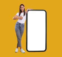 Cool Mobile Offer. Happy casual lady leaning on huge cellphone with empty white screen, showing thumb up, recommending great new app or website for cell phone, offering space for your ad, mock up photo