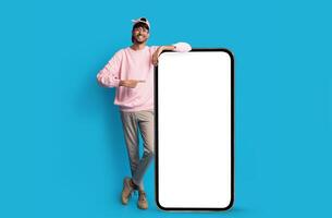 Stylish indian guy standing by huge smartphone with blank screen, mockup, pointing at advertisement, smiling millennial man recommending newest mobile application, blue studio background, copy space photo