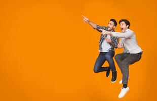 Overjoyed guys pointing at free space and jumping over orange studio background photo