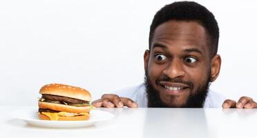 Funny Hungry Black Man Looking At Burger On Table Posing Over White Studio Background. Nutrition And Dieting, Cheat Meal And Unhealthy Junk Food, Overeating Habit Concept. Panorama photo