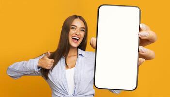 Recommendation. Portrait of excited lady holding big smartphone with white blank screen in hand, showing close up to camera making thumb up gesture. Gadget with empty free space for mock up, banner photo