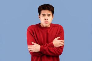 It's So Cold Here. Portrait of sad sick asian young man hugging himself to warm up, ill guy wearing sweater shivering and trembling because of frosty weather, isolated over blue studio background photo
