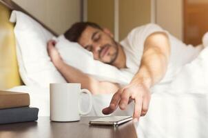 Sleepy guy waking up early after hearing alarm clock signal on smartphone on monday morning, reaching for ringing mobile phone with closed eyes, copy space photo