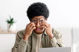 Tired black guy rubbing his eyes while studying online on laptop at home, panorama. African American male student exhausted from looking at screen, doing assignments or communicating on web photo