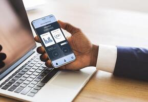 African american businessman hand holding mobile phone with trading app over laptop, creative image. Unrecognizable black manager using financial application on smartphone while working in office photo