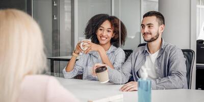 Coffee break in modern office during coronavirus outbreak and social distancing. Multiracial colleagues with cups of hot drink talk through protective glass at table with antiseptic, empty space photo