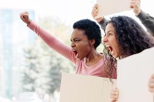 Side view of multiracial young ladies with empty placards chanting slogans and raising fists up, panorama with copy space. International group of young people students having strike on the street photo