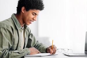 Web-based training concept. Black male student writing in notebook in front of laptop at home, empty space. Positive African American teenager studying remotely, talking to his tutor on internet photo
