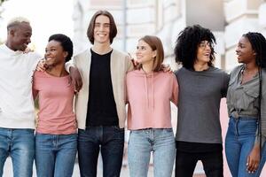 Friendship, community, team concept. Multiracial group of happy millennial men and women posing on the street, standing in a row, embracing and cheerfully smiling at camera, full length shot, panorama photo