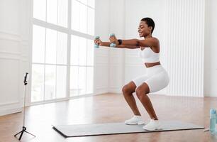 Positive black lady exercising and squatting with two dumbbells in front of cellphone on tripod at home, smiling fitness blogger recording for her followers having personal virtual training photo