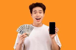 Surprised asian guy holding mobile phone with black empty screen and fan of cash money in dollar banknotes in hand, posing isolated on orange background. People and finance concept. Mock up copy space photo