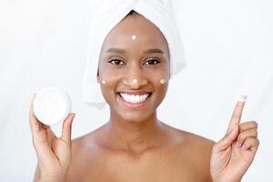 Beauty lessons and facial skin care at home during covid-19 quarantine. Millennial smiling african american lady blogger showing moisturizer cream for face in morning at home and looking in mirror photo