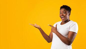 Excited Plus-Size Black Woman Showing Invisible Object Pointing Fingers Aside Advertising Your Text Posing Standing On Yellow Studio Background, Smiling To Camera. Blank Space, Panorama photo