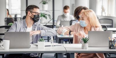 Modern contactless high five at work after lockdown and new normal. Happy millennial man and woman in protective masks touch their elbows at workplace with laptops and antiseptics in office photo