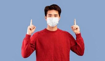 Look There. Young asian man in medical face mask pointing his fingers up at copy space above his head, isolated over studio background wall, banner. Man showing place for important information photo