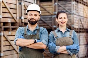 Workers in uniforms work in warehouse for sale and distribution of organic fruits. Confident smiling guy and woman with crossed arms on chest look at camera in storage, on wooden boxes background photo