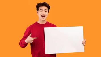Place For Your Ad. Portrait of smiling asian guy holding empty blank board isolated on orange studio background. Happy guy standing with white square paper for template and pointing at it photo