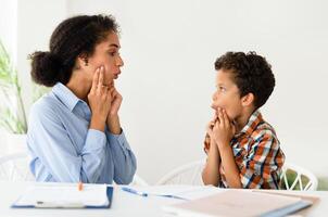Speech Therapy Session. Woman working on kid boy pronunciation sitting at desk indoors. Professional speech therapist lady helping little patient with stuttering issue. Logopedic correction photo
