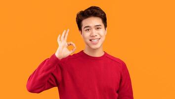 Everything Is Okay. Portrait of positive smiling asian guy doing approval ok gesture with fingers, showing that he is fine, isolated over orange studio background. Confident man is cool photo