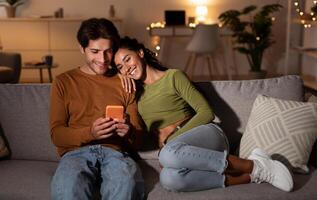 Young Couple Using Phone Browsing Internet Relaxing Sitting On Sofa At Home At Night. Husband And Wife Texting Via Cellphone Or Checking New Mobile Application On Smartphone Indoor. Modern Gadgets photo