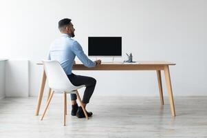 Rear back view portrait of Middle Eastern businessman looking at blank empty computer monitor sitting at desk at workplace, watching webinar or having online web call, free copy space banner photo