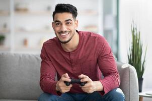 Emotional middle-eastern guy playing game with joystick at home, copy space. Happy millennial arab man enjoying weekend, sitting on couch in living room with joystick, gaming photo