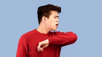 Stop Virus And Germs Spreading Concept. Closeup portrait of asian guy in sweater showing correct way of sneezing, coughing in his elbow isolated over studio backgound, banner, panorama, copy space photo