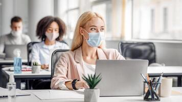 Modern coworking office worker after returning from covid quarantine to office. Millennial woman in protective mask with smart watch sitting at table with laptop, looks at window and creating idea, empty space photo