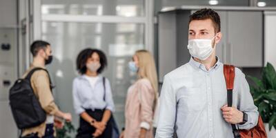 New normal and returning to work after coronavirus epidemic. Young guy with backpack in protective mask looks to the side in interior of office, colleagues are standing behind, blurred background, free space, indoors photo