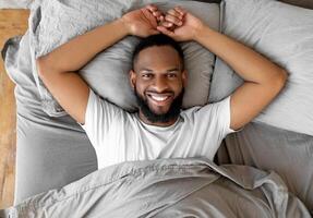 Overhead above top view portrait of relaxed smiling African American man lying on gray bedsheets at home after waking up, feeling good, peaceful young black guy resting in bedroom looking at camera photo