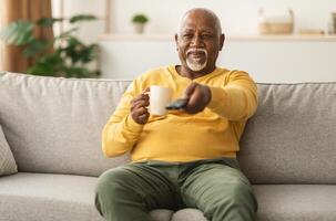 Contented Senior African American Man Watching TV Pointing Television Remote Control To Camera Switching Channels Sitting On Couch At Home. Male Viewer Enjoying Movie On Weekend photo