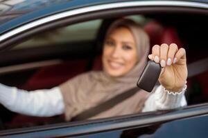 Test Drive New Car. Smiling Arabian Woman In Hijab Showing Automobile Keys Out Of Window, Sitting In Vehicle. Confident Middle East Lady Posing In Driver's Seat, Blurred Background, Selective Focus photo