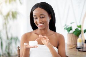 Home spa and permanent facial during covid-19 lockdown. Cheerful beautiful millennial african american female applying face cream to moisturize skin in bedroom interior in morning, copy space photo