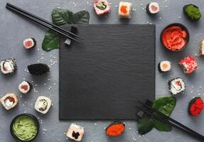 Top view on traditional japanese empty square black slate framed with sushi, ginger, soy sauce, wasabi bowls, green leaves and chopsticks on rustic grey background, copy space photo