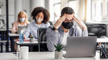 New normal and brainstorming while working in office during covid-19 outbreak. Millennial man in protective mask rests his head on hands, sitting at table and looking at laptop at table with antiseptic photo