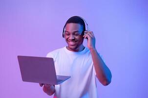 Black Gamer Man In Wireless Headset Playing Games On Laptop, Looking At Computer Screen While Standing In Vivid Neon Light Over Purple Studio Background, Guy Enjoying Cyber Gaming, Copy Space photo