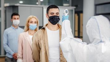 New normal and coronavirus disease control. Medic in protective uniform with infrared thermometer to measure temperature of workers in face masks, who came to work in office in morning, copy space photo