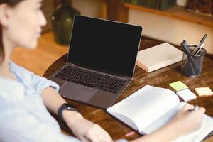 Online Course Concept. Over the shoulder view of female student sitting at table and writing in notebook, using laptop with black empty screen for mock up template, watching lecture or webinar photo