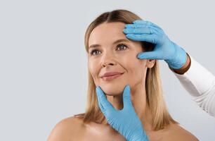 Plastic surgery or face lifting for middle aged woman. Doctor hands wearing blue medical gloves holding head of female patient, checking skin before procedure, isolated on grey background, copy space photo