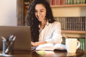 Workspace Concept. Portrait of smiling curly woman using and working on computer, typing on laptop keyboard. Young lady sitting at desk with open notebook and a cup of coffee, blurred background photo