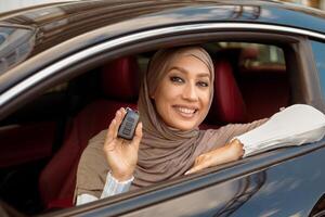 Test Drive New Car. Smiling Arabian Woman In Hijab Holding And Showing Automobile Keys Out Of Window, Sitting In Vehicle. Confident Middle East Buyer Posing In Driver's Seat, Selective Focus photo