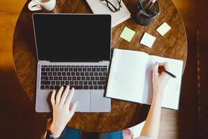 Elearning Concept. Above top view of woman sitting at desk and writing in notebook, taking notes, using laptop with black empty screen for mock up template, typing on keyboard, working on project photo