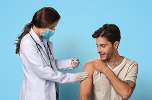 Vaccination Concept. Female Doctor With Syringe Making Vaccine Injection To Male Patient's Arm, Young Smiling Man Getting Vaccinated Against Covid-19 While Sitting Over Blue Background, Free Space photo