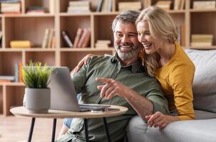 Digital Entertainment. Cheerful Middle Aged Couple Using Laptop At Home Together, Smiling Spouses Relaxing In Living Room With Computer, Watching Movies Or Purchasing Online, Man Pointing At Screen photo