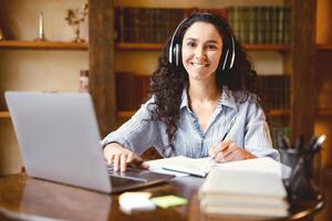 Telecommuting. Portrait of smiling woman in wireless headset sitting at desk, using laptop and writing in notebook, looking at camera, watching lecture or webinar, studying or working online at home photo