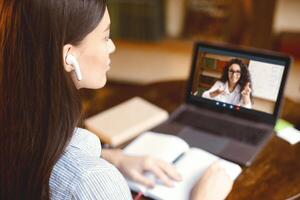 Distance Education Concept. Over the shoulder view of student wearing earbuds, sitting at desk with open notebook, using laptop, having chat with colleauge or watching online class with teacher photo