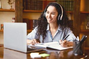 Distance Education. Smiling woman in wireless headset sitting at desk, using laptop and writing in notebook, taking notes, watching tutorial, lecture or webinar, studying online at home. E-learning photo