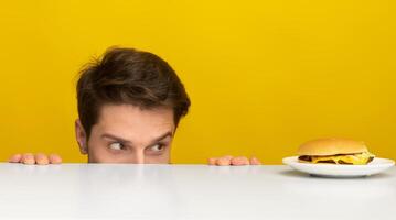 Funny Hungry Man Looking At Burger On Plate, Peeking Out Table Posing Over Yellow Orange Studio Background. Dieting, Cheat Meal And Unhealthy Junk Food, Overeating Habit Concept. Panorama Banner photo