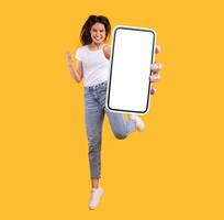 Yes. Full body length portrait of joyful woman jumping up, making winner gesture with clenched fist and showing cellphone with free empty space for mobile app on white screen, orange studio background photo
